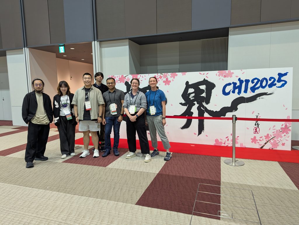 Picture of seven people standing in front of a CHI2025 sign/artwork. 